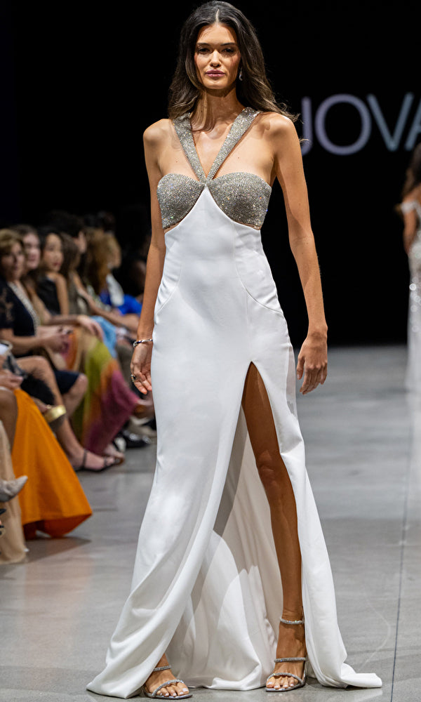 A model walks the runway in Jovani's Long Prom Dress 36857, a sleeveless white jersey fitted gown with a high slit and crystal-embellished silver straps and bodice, as audience members watch from the sides.